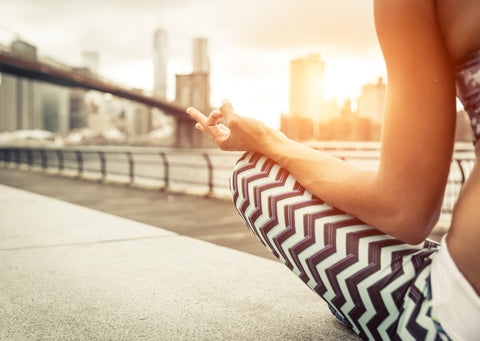 Woman in chevron leggings meditating with a mudra hand gesture by a city waterfront at sunset
