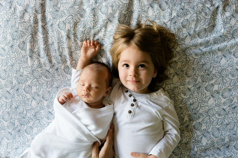 Young child lying beside a sleeping swaddled newborn baby on a floral bedsheet