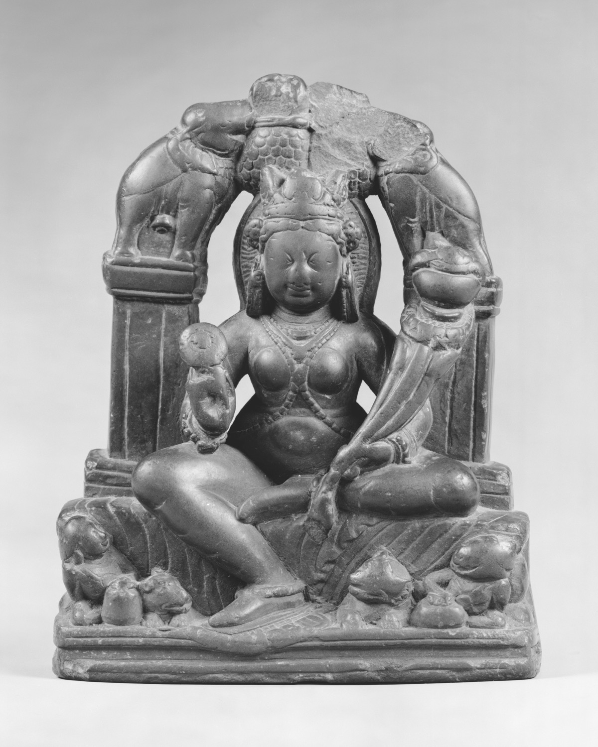 A dark stone figure of Lakshmi seated on a carved base, with two elephants arched over her head