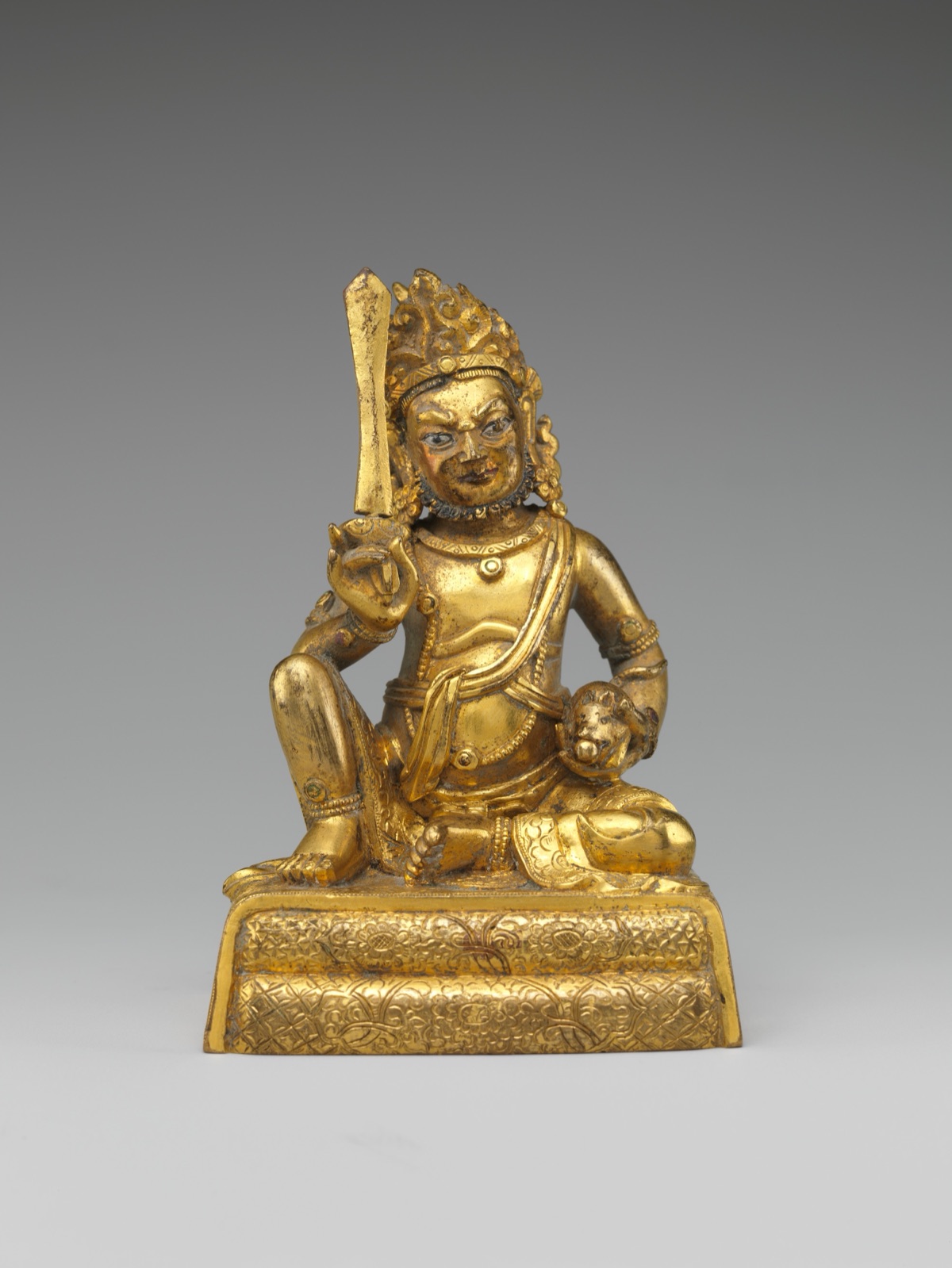 A gold-coloured brass statue of a seated Kubera raising a flat, blade-shaped object, a small animal resting in his other hand