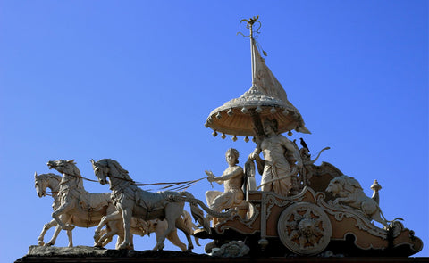 Stone statue of Krishna and Arjuna in a horse-drawn chariot under a canopy, set against a blue sky