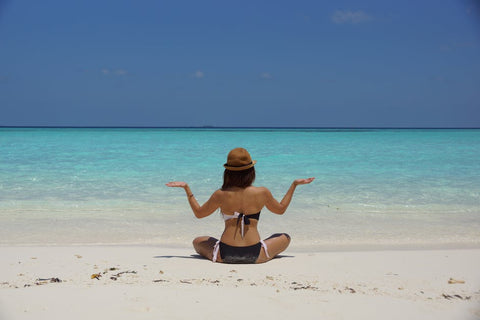 Woman in a bikini and straw hat sitting cross-legged on a white beach facing turquoise sea