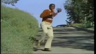 Animated GIF of a man in an orange sweater dancing down a sunny hillside road