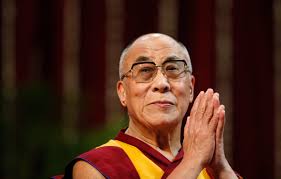 The Dalai Lama in maroon and yellow robes and glasses, palms pressed together