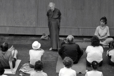 Black-and-white photo of an elderly woman teacher in a dark robe standing before seated yoga students on mats