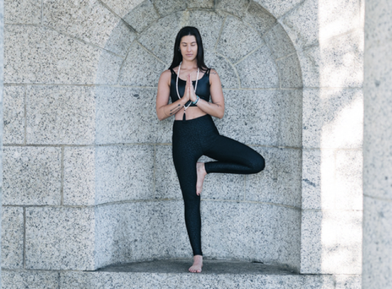 Woman in Tree Pose (Vrksasana) with hands in prayer position, standing in a stone archway