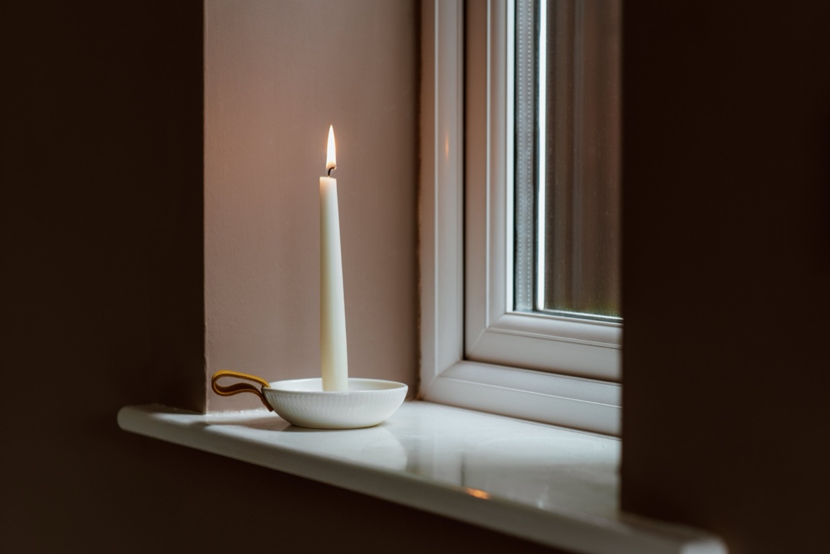 A single lit white candle in a small white holder on a windowsill in a dim room