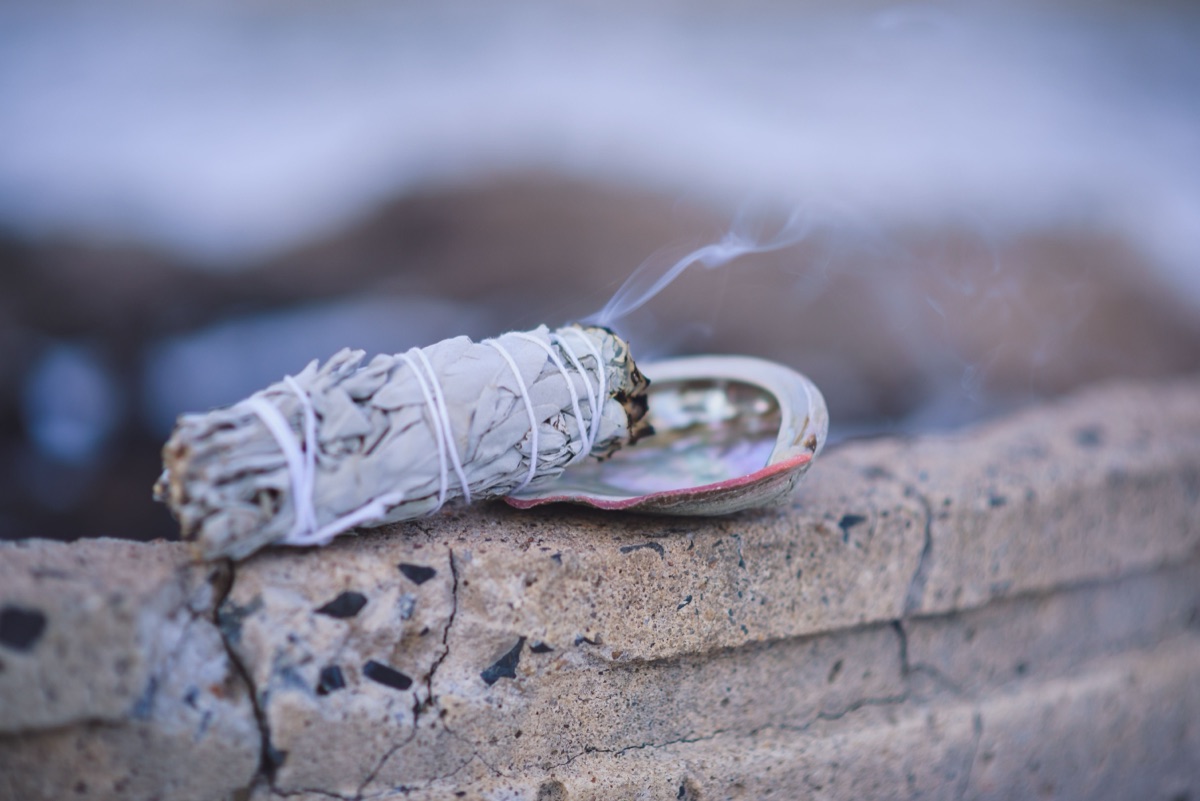 A white sage bundle wrapped in white thread, smoke rising from its tip, resting on a shell on a stone ledge