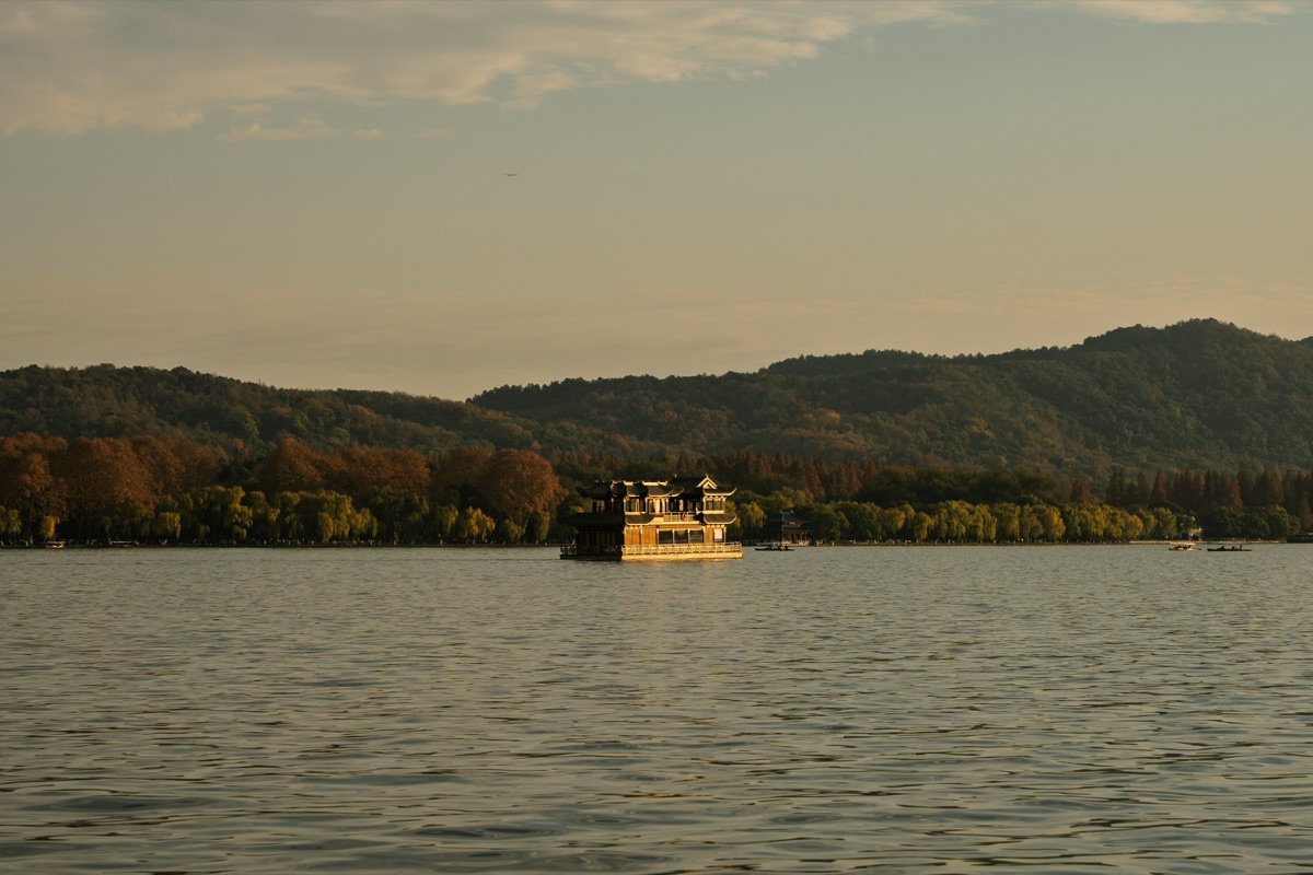 Calm water of West Lake in Hangzhou at golden hour, a traditional boat on the water and a low line of wooded hills behind