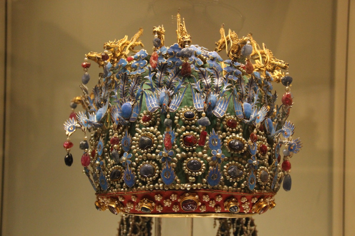 A Ming empress's crown in a museum case, covered in blue phoenix ornaments, pearls and red and blue gems, with gold dragons along the top
