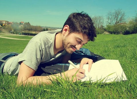 Smiling young man lying on grass in a park, writing in an open notebook