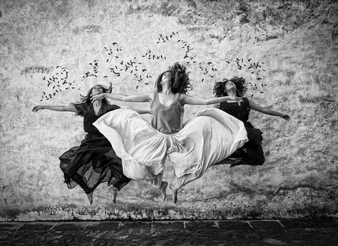 Black-and-white photo of three women in dresses leaping mid-air in front of a textured wall