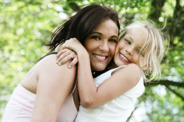 Smiling mother and young blonde daughter hugging outdoors under green trees