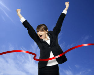 Businesswoman in a black suit throwing her arms up as she breaks a red finish-line ribbon