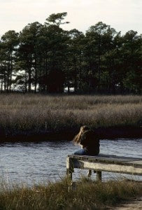 Woman sitting alone at the end of a wooden dock by a marsh, facing tall pine trees