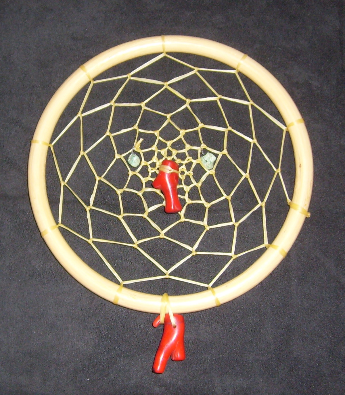 A pale wooden hoop filled with a web of yellow synthetic sinew, two small turquoise stones and red coral pendants at the centre and bottom