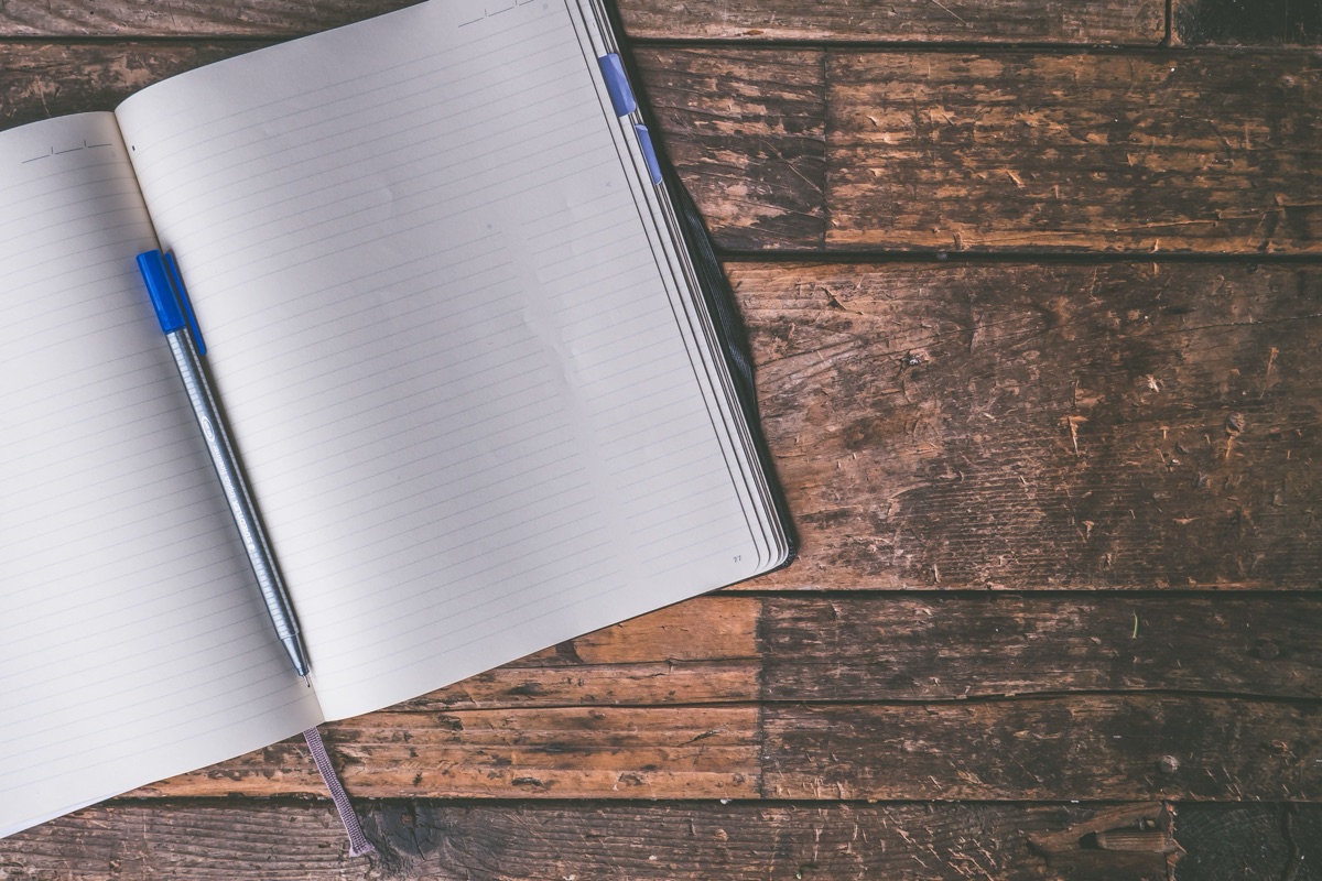 An open lined notebook with a blue-capped pen lying across its pages on weathered wooden boards