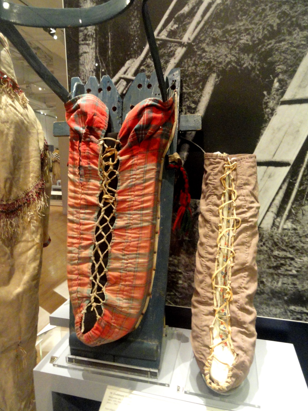 An Ojibway cradleboard with a red plaid laced wrap and a curved bow over the top, beside a laced moss bag in a museum case