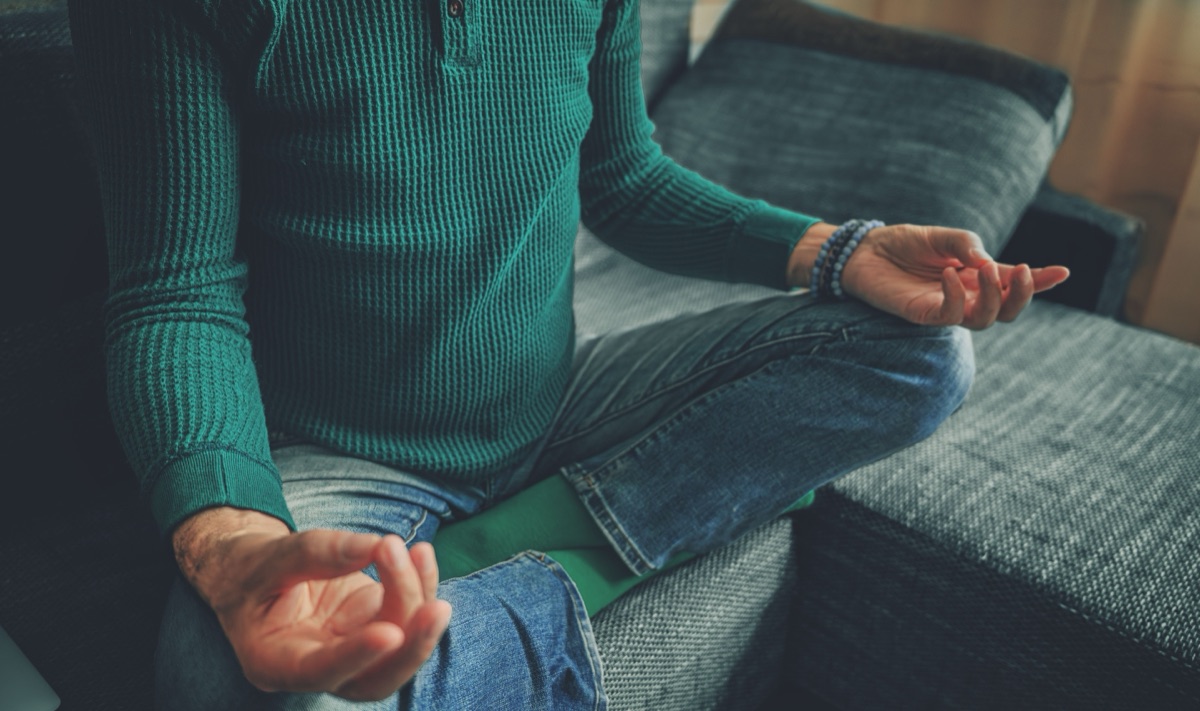 A person sitting cross-legged on a grey couch, each hand resting on a knee with thumb and finger touching, a bead bracelet on one wrist
