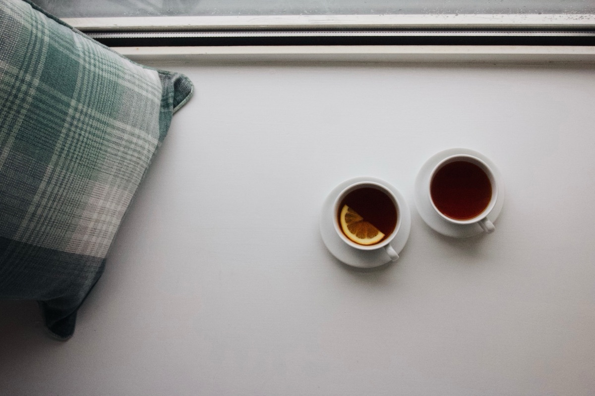 Two cups of tea on saucers side by side on a white windowsill, next to a green plaid cushion