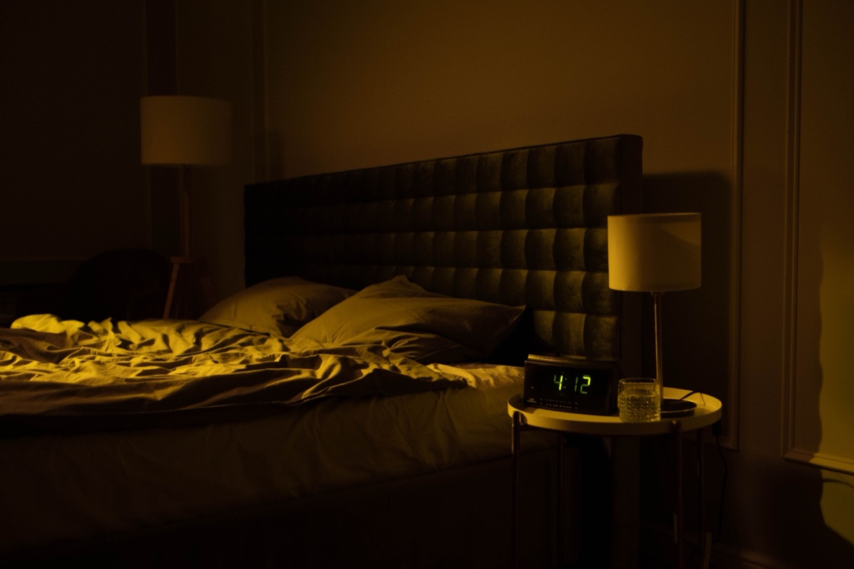 An empty unmade bed in a dim bedroom, with a lamp and a digital clock reading 4:12 on the bedside table