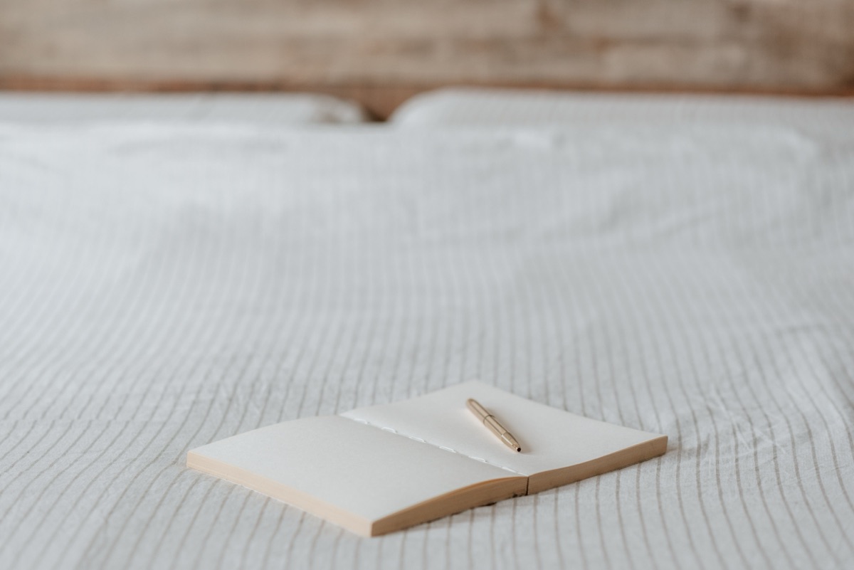 An open blank notebook with a gold-coloured pen on its right-hand page, lying on a striped white bedsheet