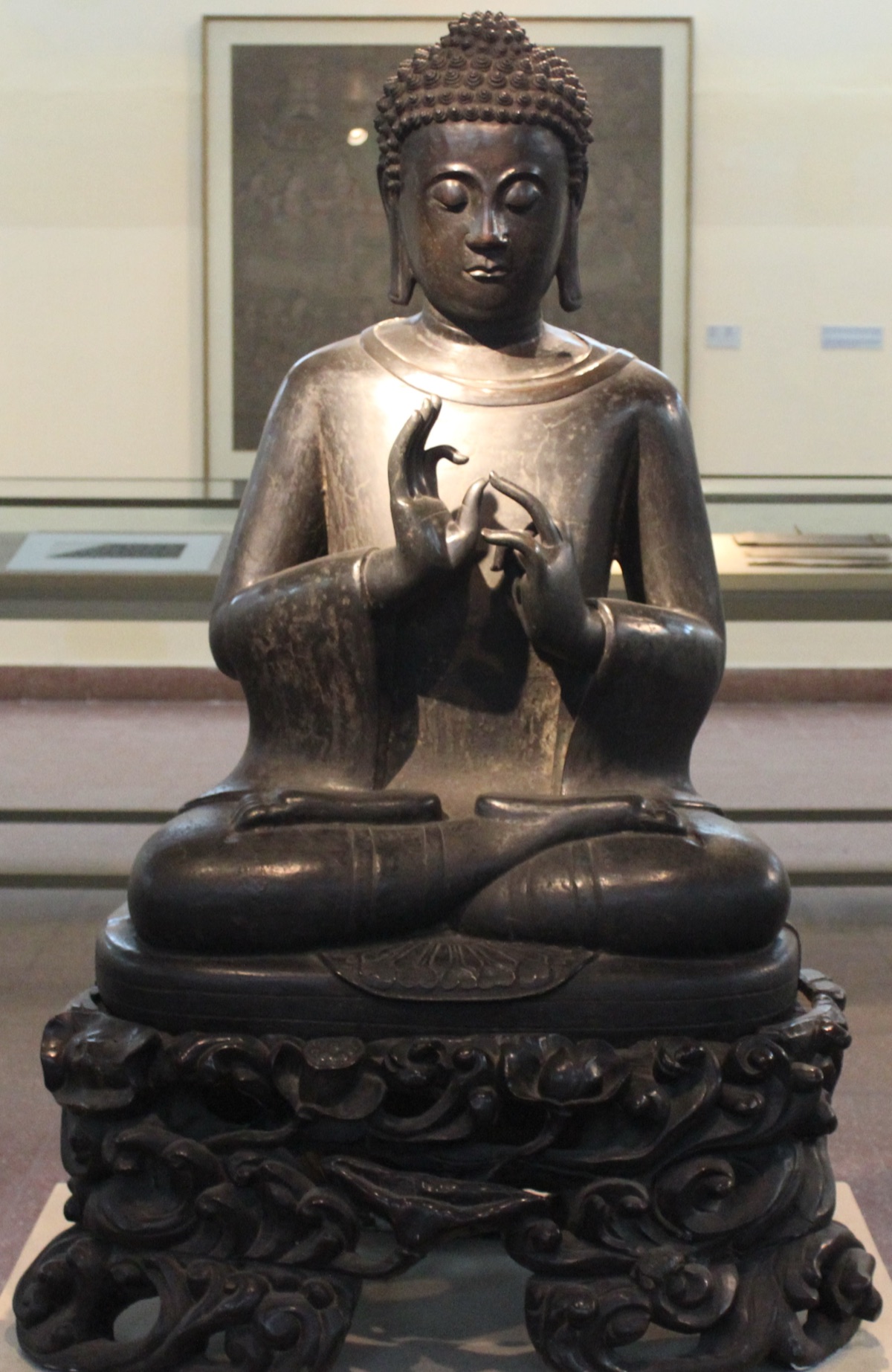 A dark stone seated Buddha in a museum gallery, both hands at the chest, the raised right hand joining thumb and index finger in a circle