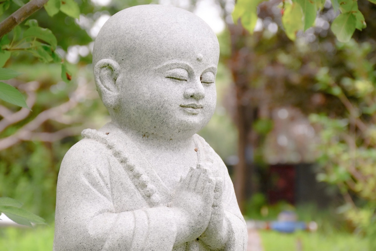 A granite statue of a young shaven-headed monk wearing prayer beads, eyes closed, palms pressed together at the chest