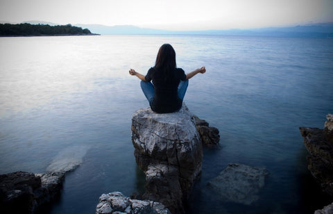 Woman meditating cross-legged on a rock at the edge of calm water