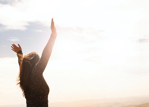 Woman seen from behind raising both arms toward a bright hazy sky