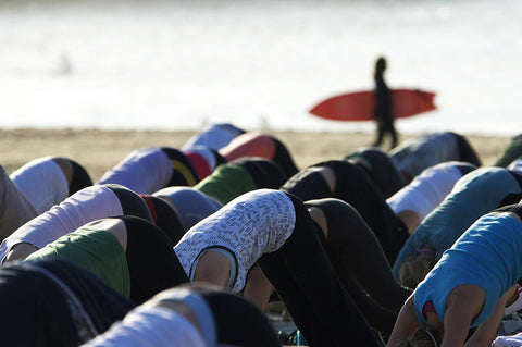 Rows of people in downward dog on a beach while a surfer walks past with a red board
