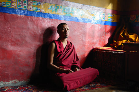 Buddhist monk in maroon robes meditating cross-legged against a red painted monastery wall